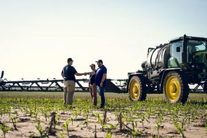 Agricultural professionals discuss farm operations beside a John Deere sprayer in a newly planted crop field.