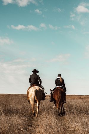 Two riders on horseback traverse through tall grass prairie against a blue sky with white clouds.