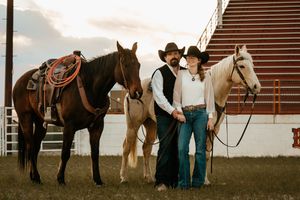 A couple in cowboy hats and western attire poses with two horses near a rodeo arena at sunset.