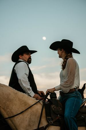 A cowboy and cowgirl on horseback wearing black hats face each other under a moonlit sky at dusk.