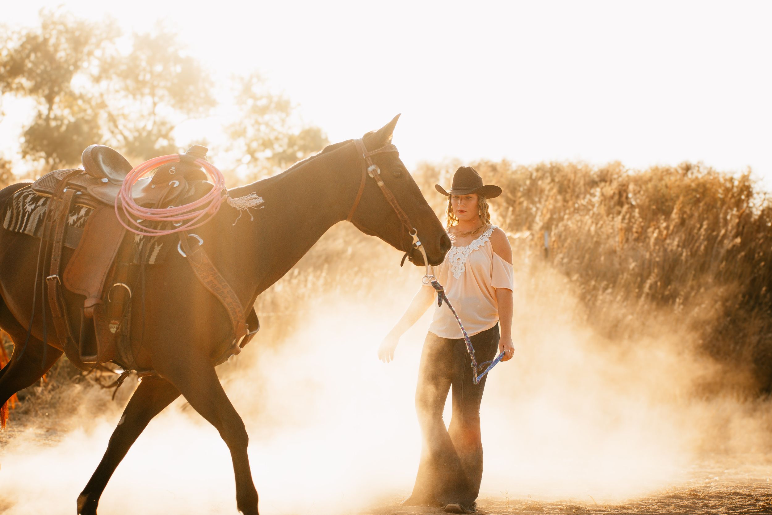 Horse and rider silhouetted against golden sunlight in dusty ranch setting.