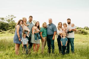 Large family group portrait smiling outdoors in a green meadow, including adults, toddlers, and babies on a sunny day.