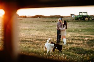 A couple embraces in a golden field at sunset with a tractor silhouetted in the background.