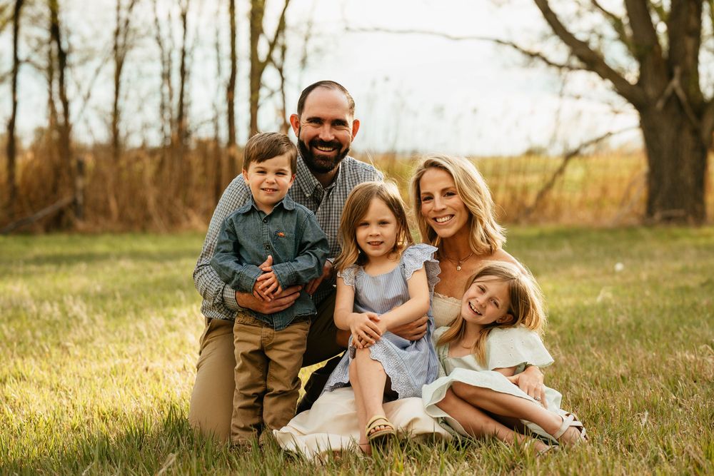 Happy family of five posing outdoors in a sunny field, parents smiling with three young children in casual attire.