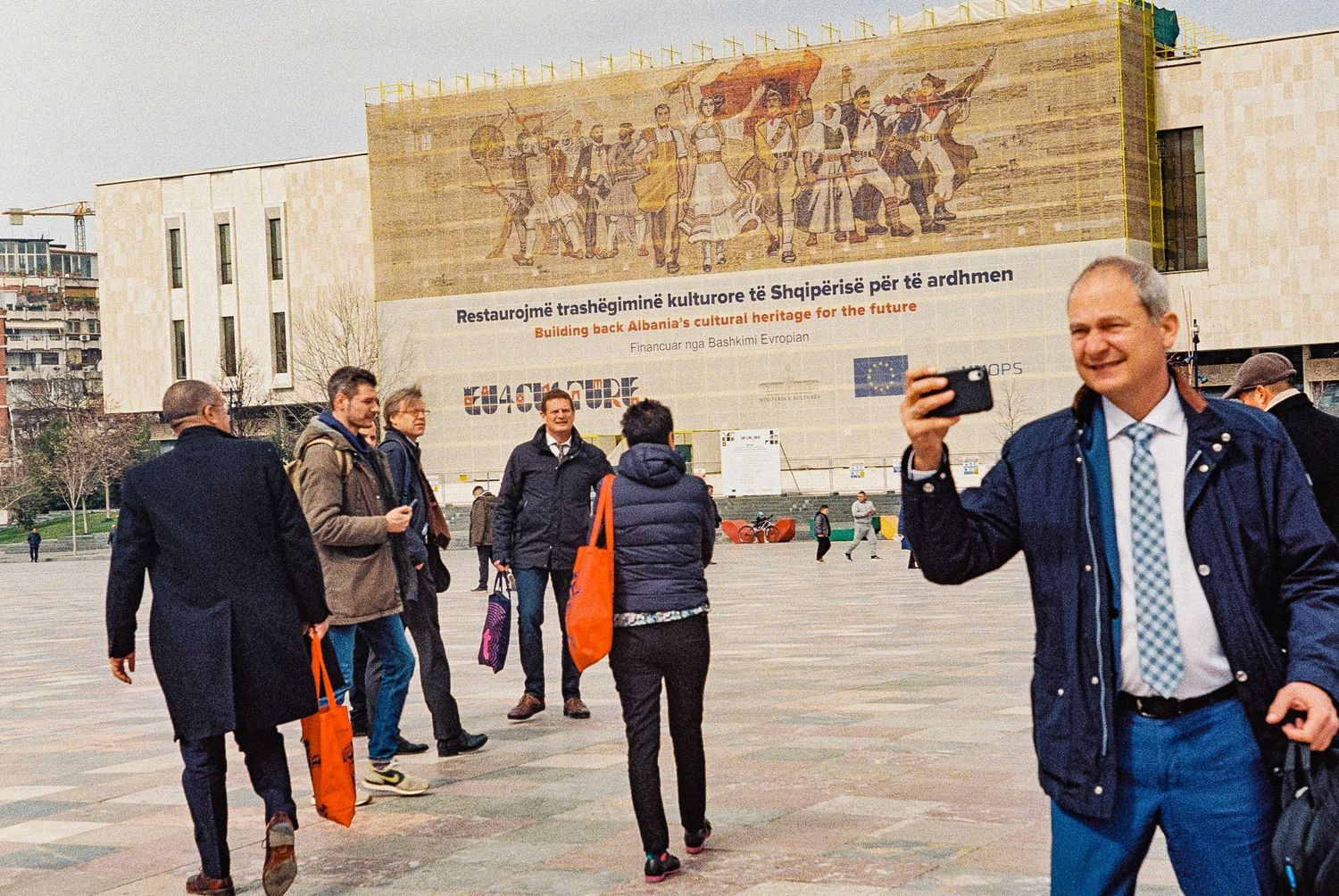 Public square with Soviet-era mural and people walking in Tirana, Albania.