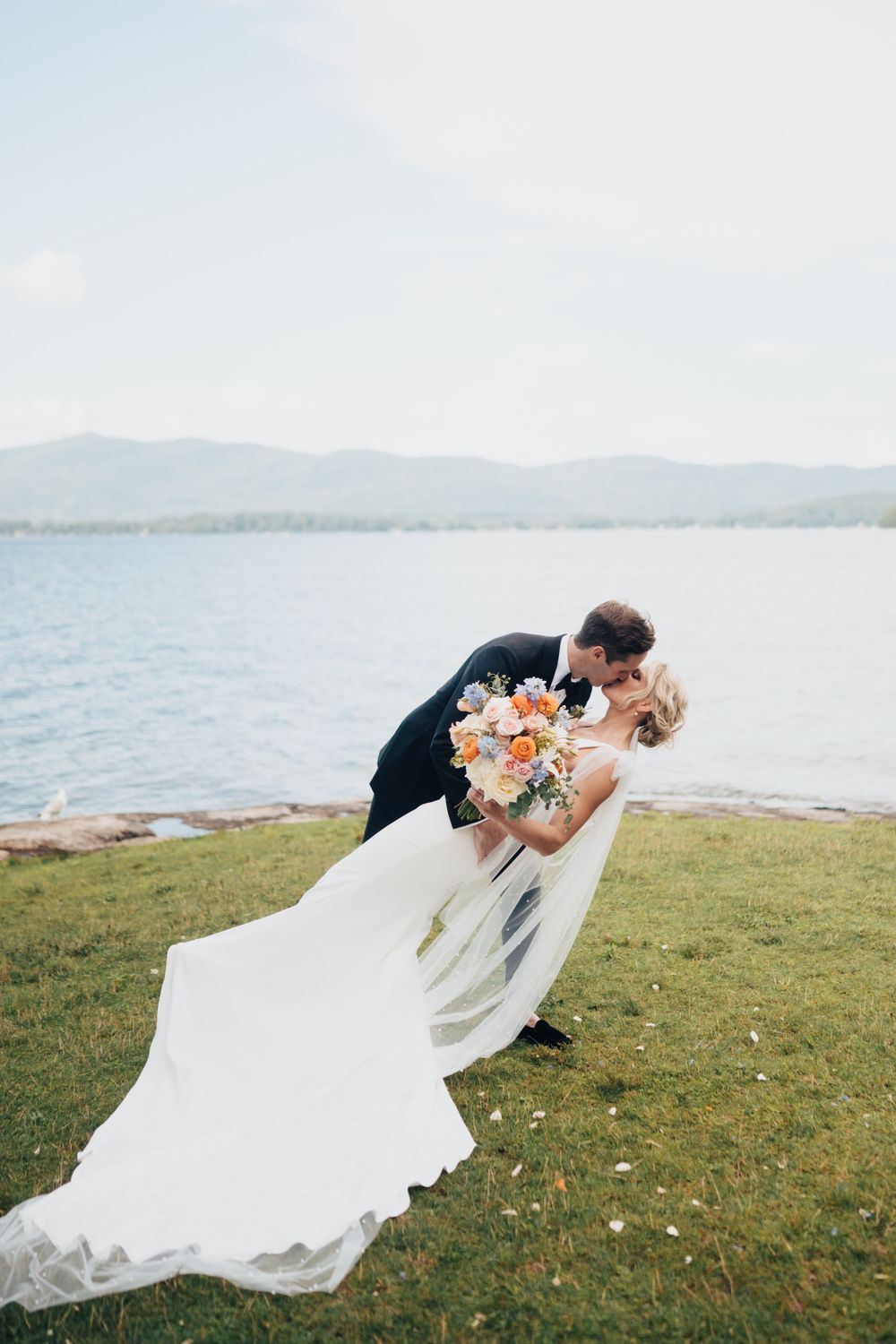 Groom dipping bride for a romantic kiss on a lakeside lawn with mountain backdrop on wedding day.