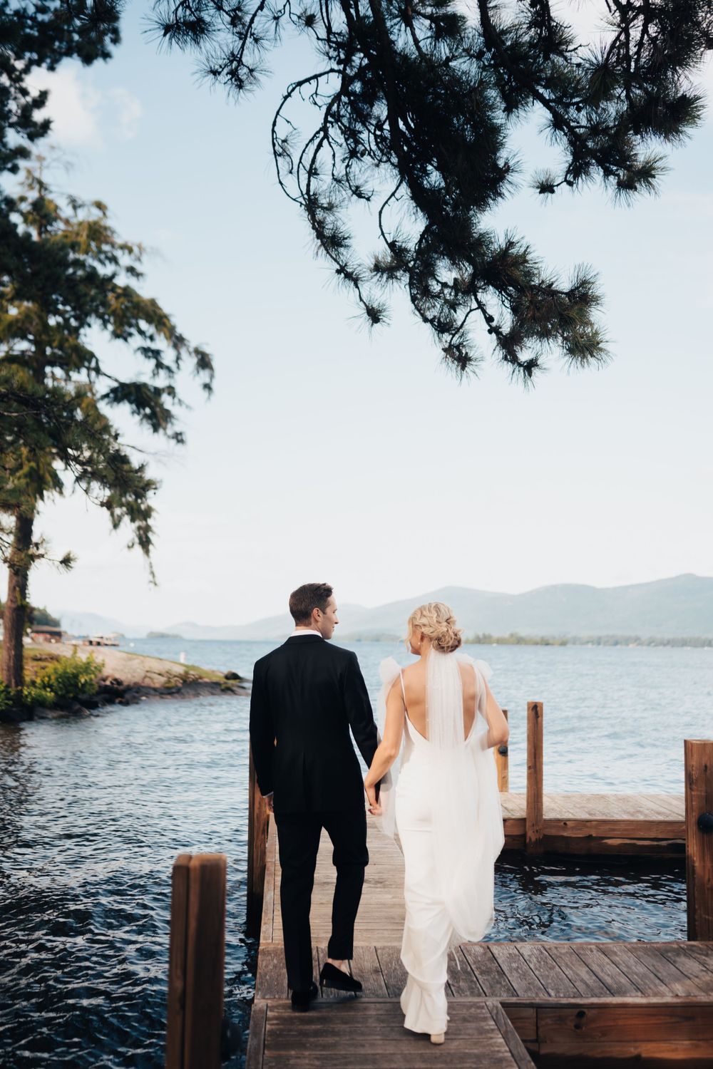 Bride and groom walk hand-in-hand on a wooden dock overlooking a serene mountain lake at sunset.