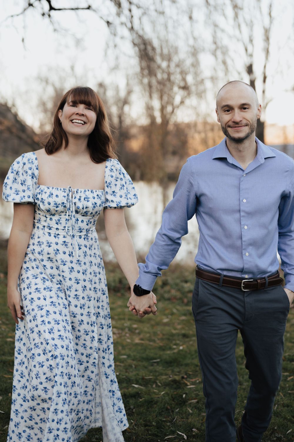 Smiling couple walks hand-in-hand through a park in casual attire during a golden hour engagement session.