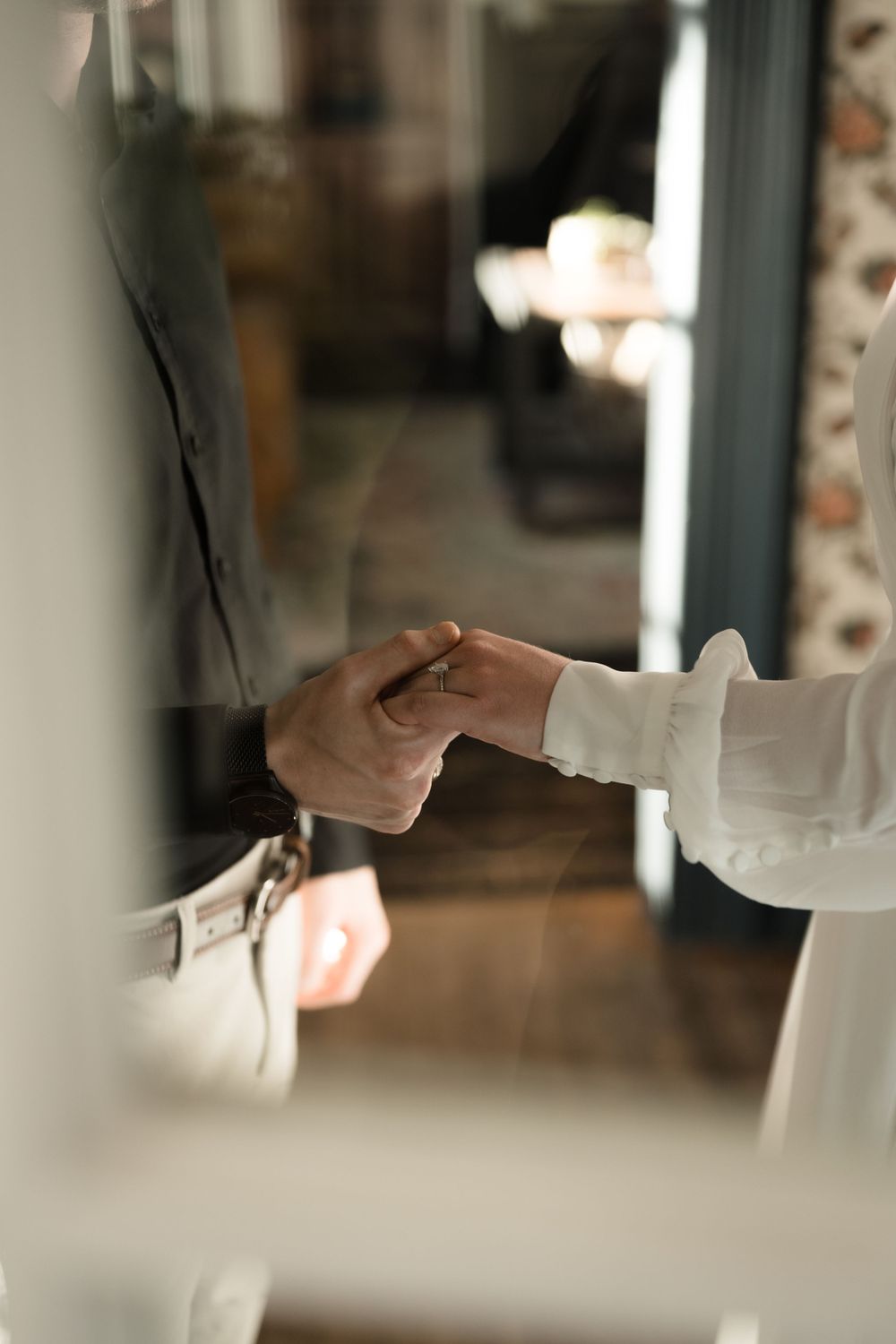 Close-up of a bride and groom tenderly holding hands in a softly lit indoor wedding setting.