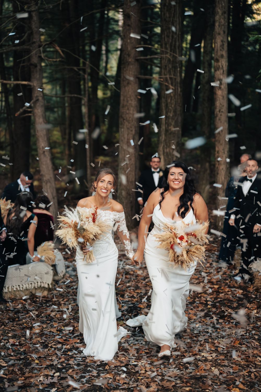 Two brides walking joyfully through a forest ceremony aisle as confetti rains down on wedding day.