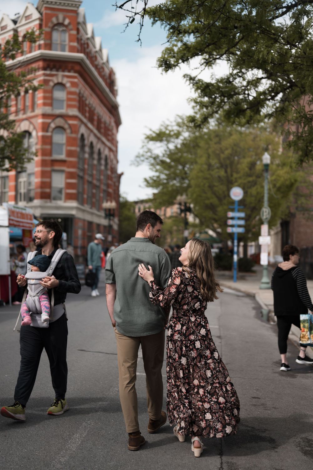 Couple walking hand in hand through a bustling city street with a historic red brick building backdrop.