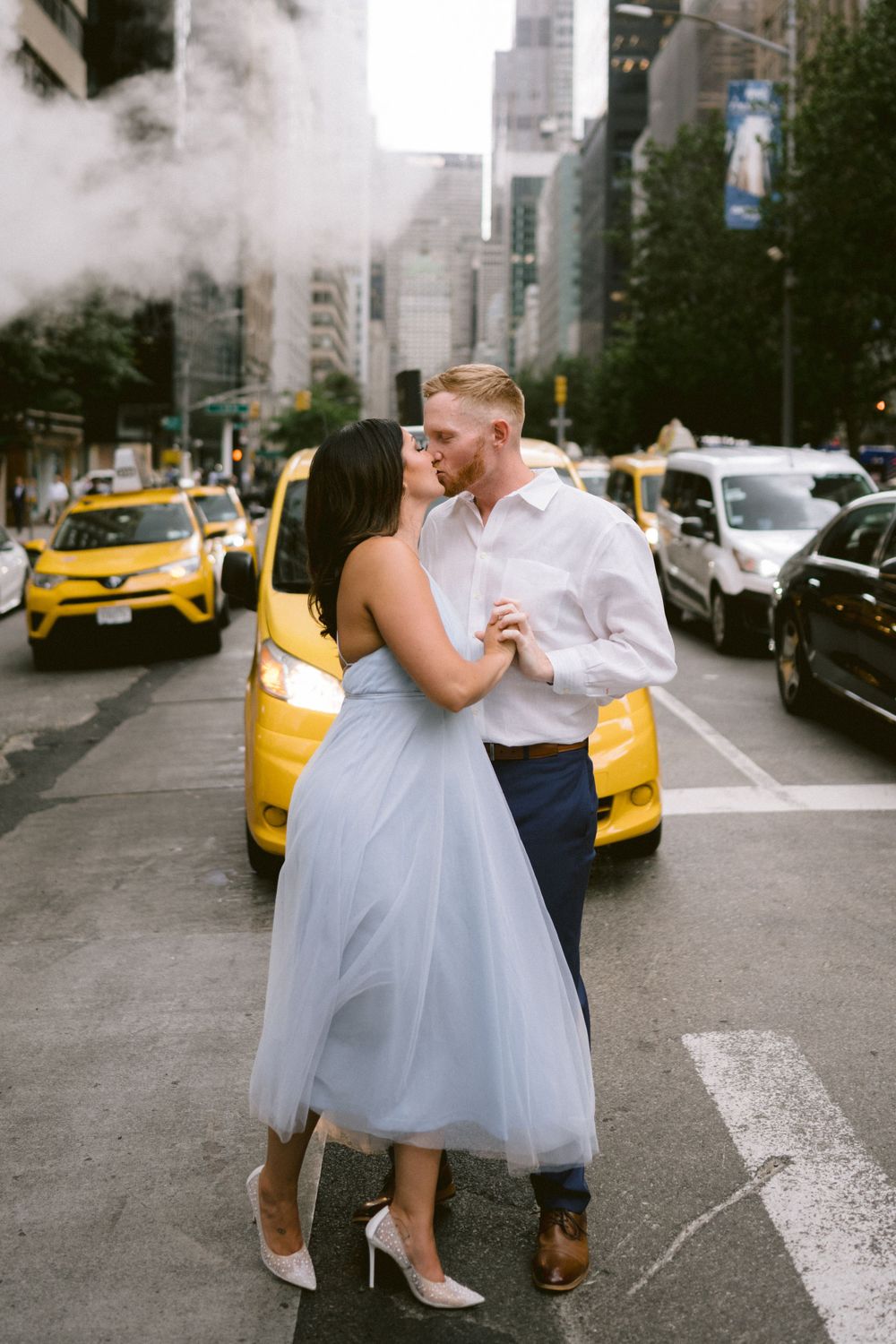 Couple shares a kiss while dancing on a New York City street with yellow taxis passing behind them.