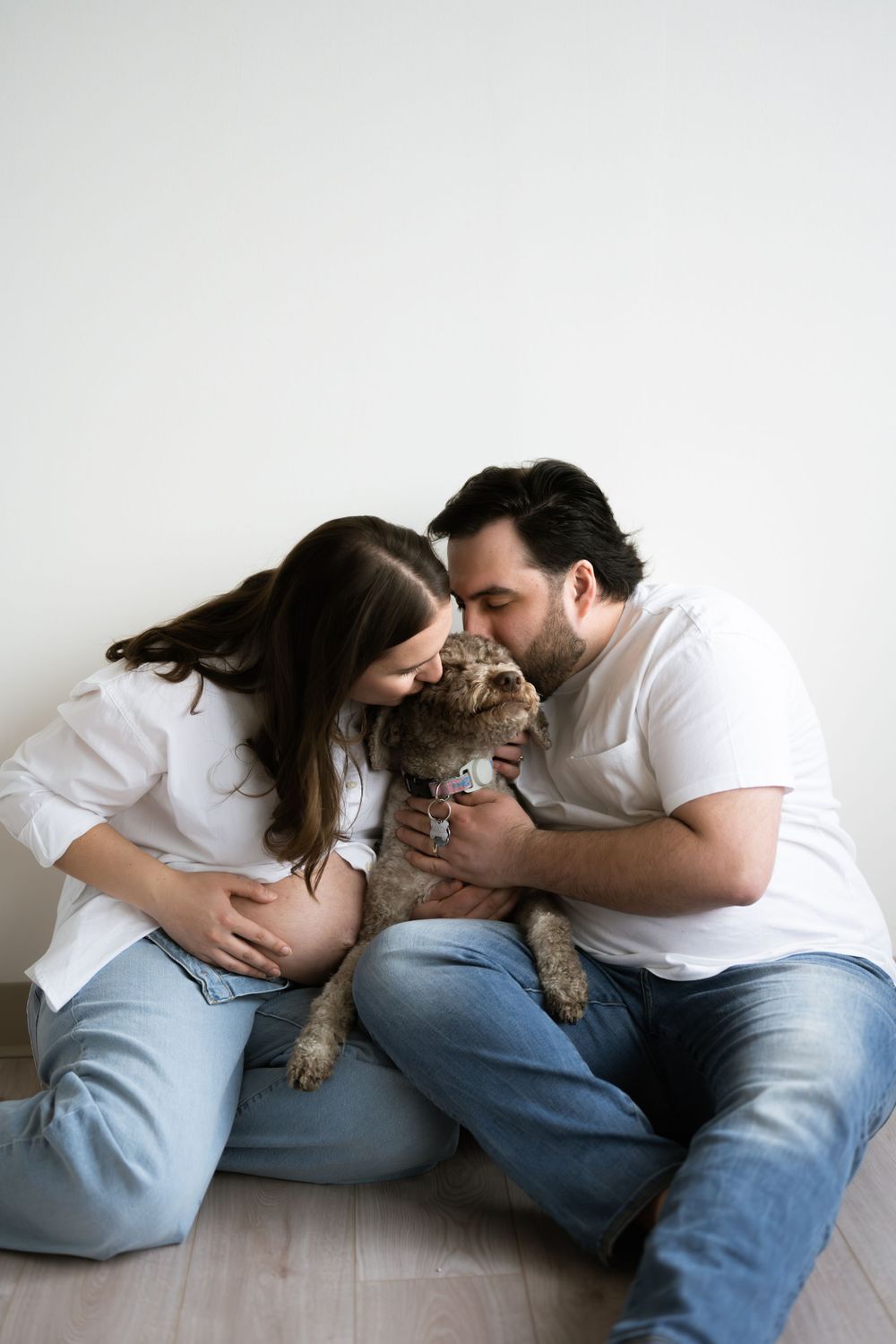 Expecting couple sitting on floor kissing their brown dog while woman cradles her pregnant belly.