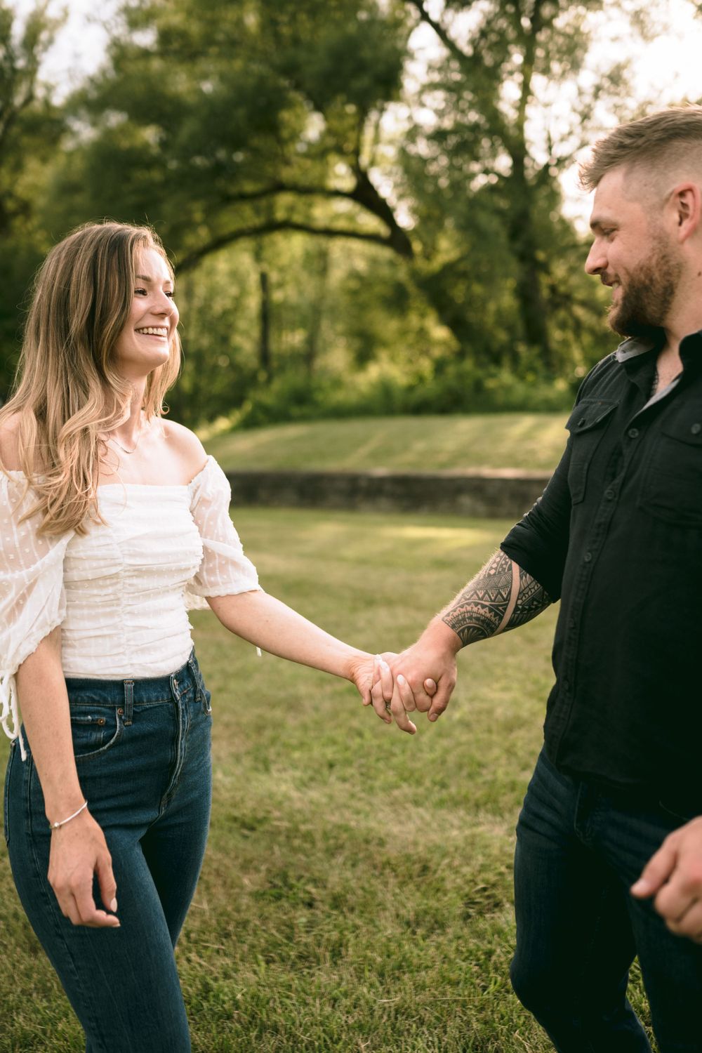 Smiling couple holds hands in a lush green park, woman in white top and jeans, man with tattooed arm.