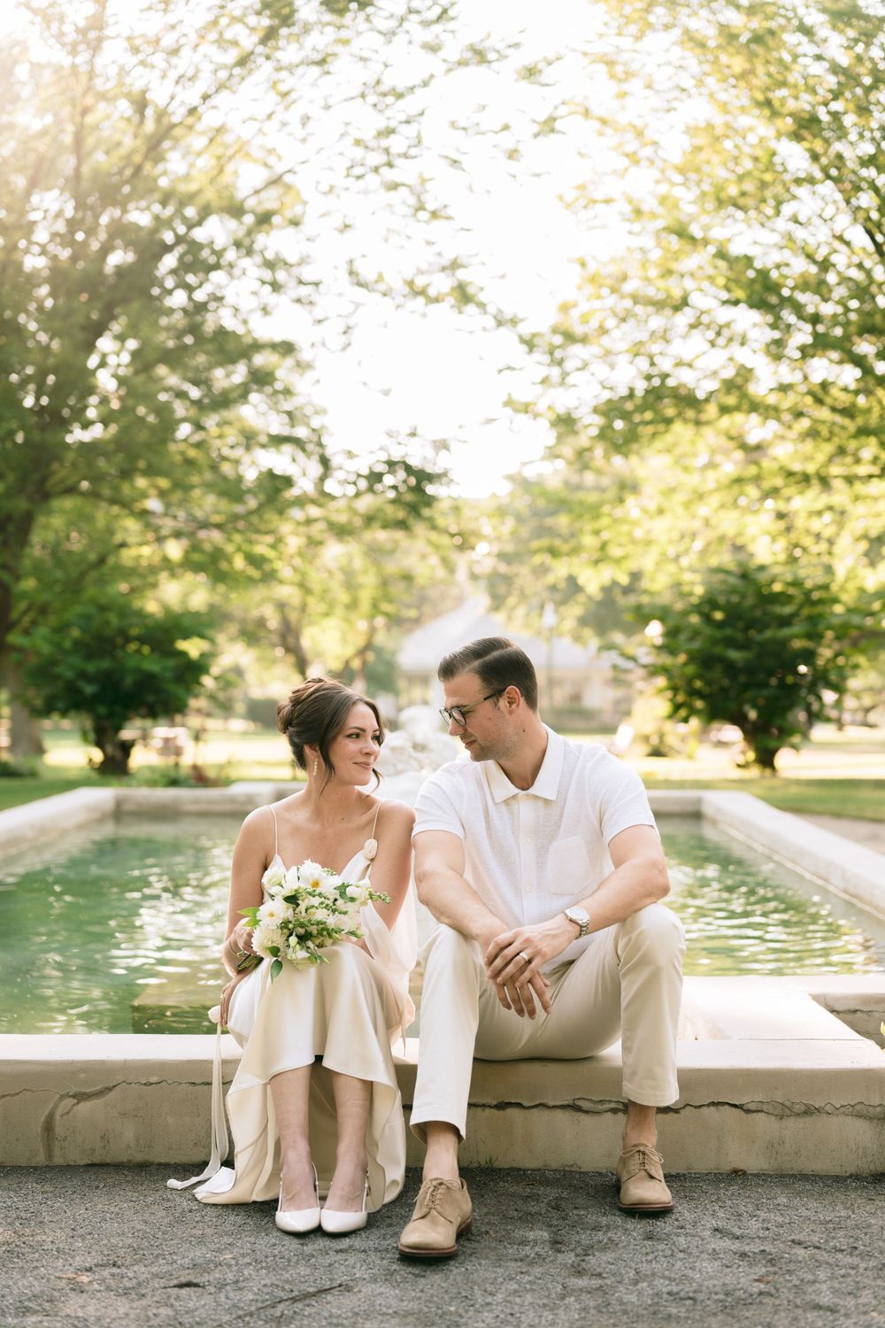 Bride and groom sit together by a sunlit fountain in a lush green park, sharing a quiet moment.