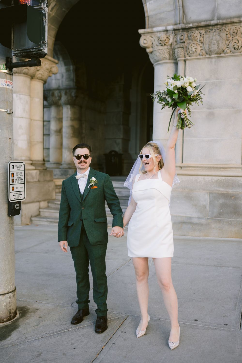 Joyful newlyweds celebrate outside a grand building, bride raising bouquet in a short white dress and sunglasses.
