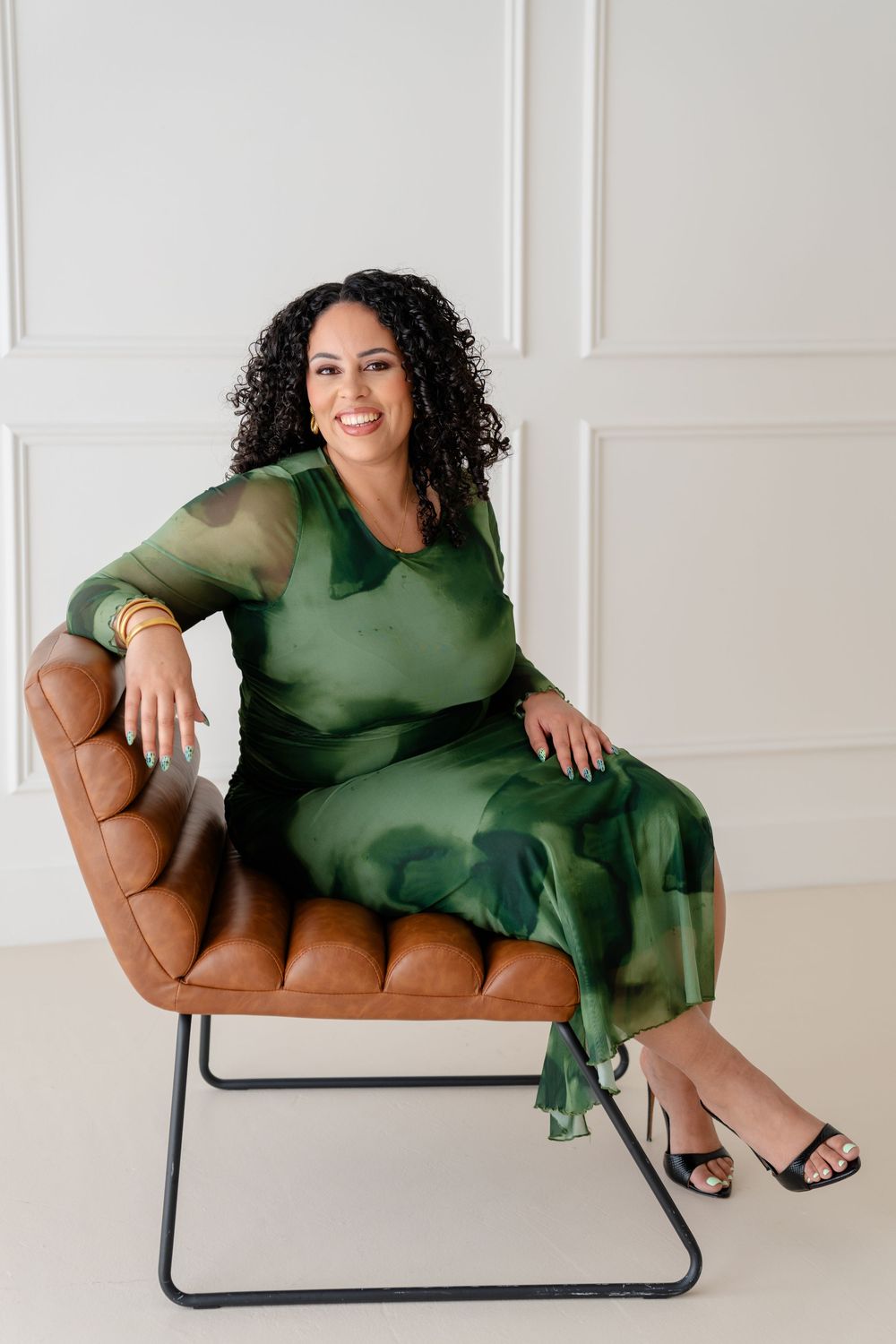 A person in a green dress lounges elegantly across a tan leather chair in a bright white studio setting.