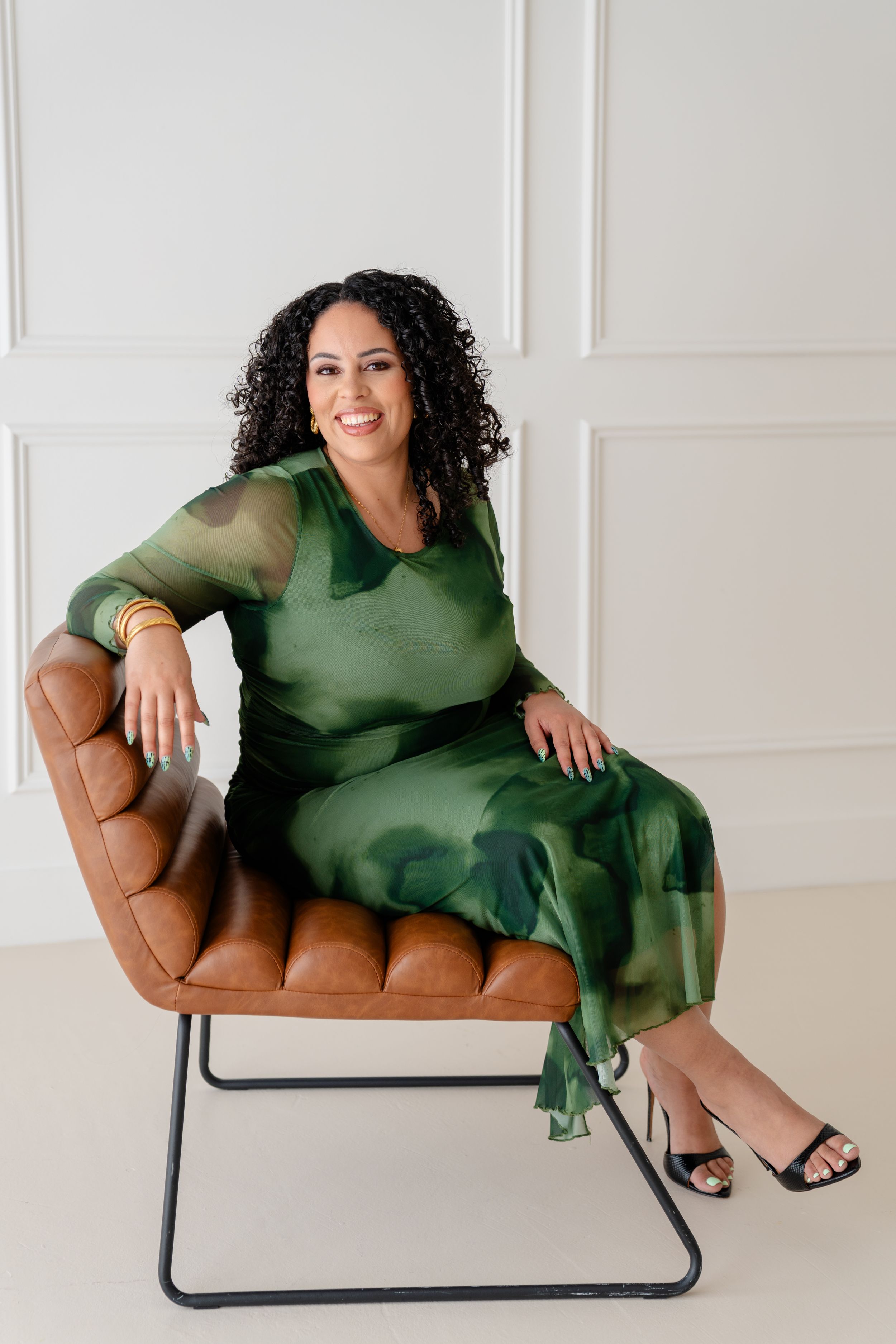 A person in a green dress lounges elegantly across a tan leather chair in a bright white studio setting.