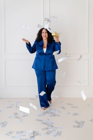 Person in royal blue business suit throws money in the air while sitting and posing playfully on white floor.
