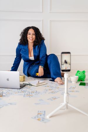 Content creator in blue suit working on floor with laptop, phone on tripod, and green frog decoration nearby.