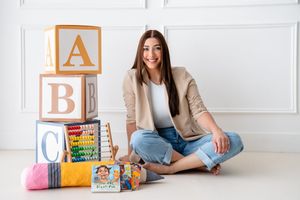 A person sits cross-legged on floor near ABC blocks and children's books wearing beige blazer and jeans in bright room.