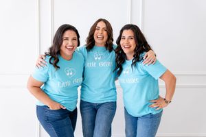 Three women wearing matching light blue t-shirts and jeans pose together against a white background.