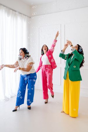 A group of dancers in colorful outfits perform joyful movements in a bright white studio space.