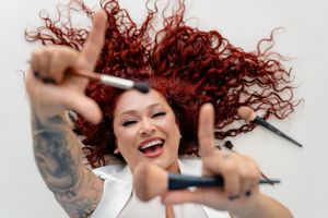 A person with vibrant curly red hair appears joyful and energetic with arms raised and tattoos visible in a candid shot.