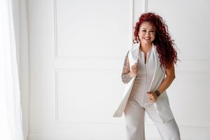 Stylish businessperson with curly red hair wearing a white pantsuit poses against bright white wall.