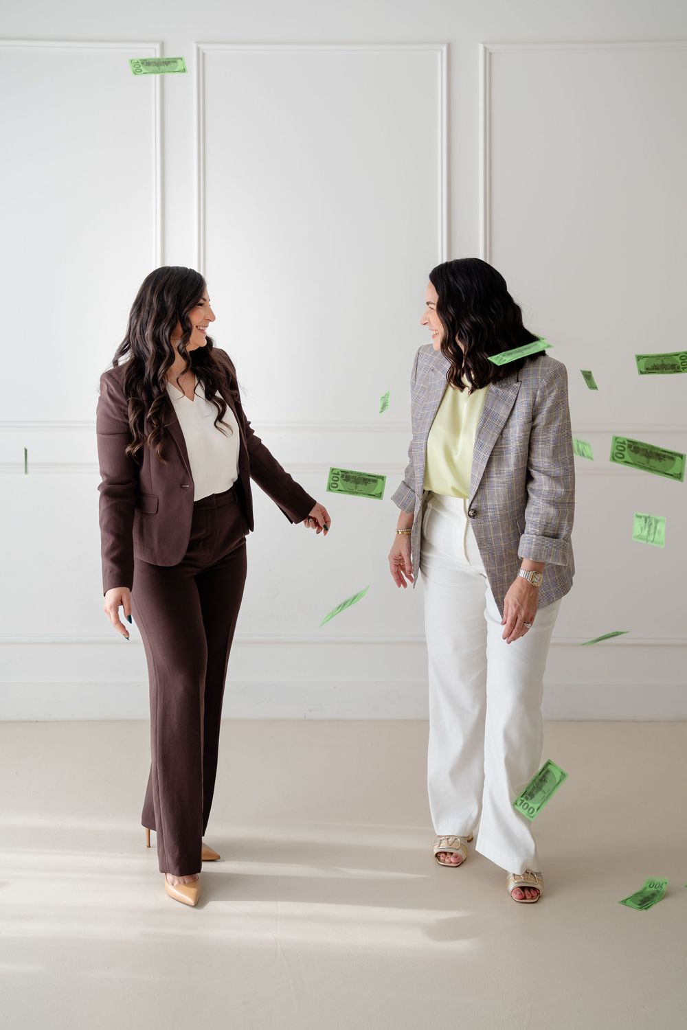 Two people in contrasting brown and white outfits interact playfully as green paper money falls around them in a bright room.