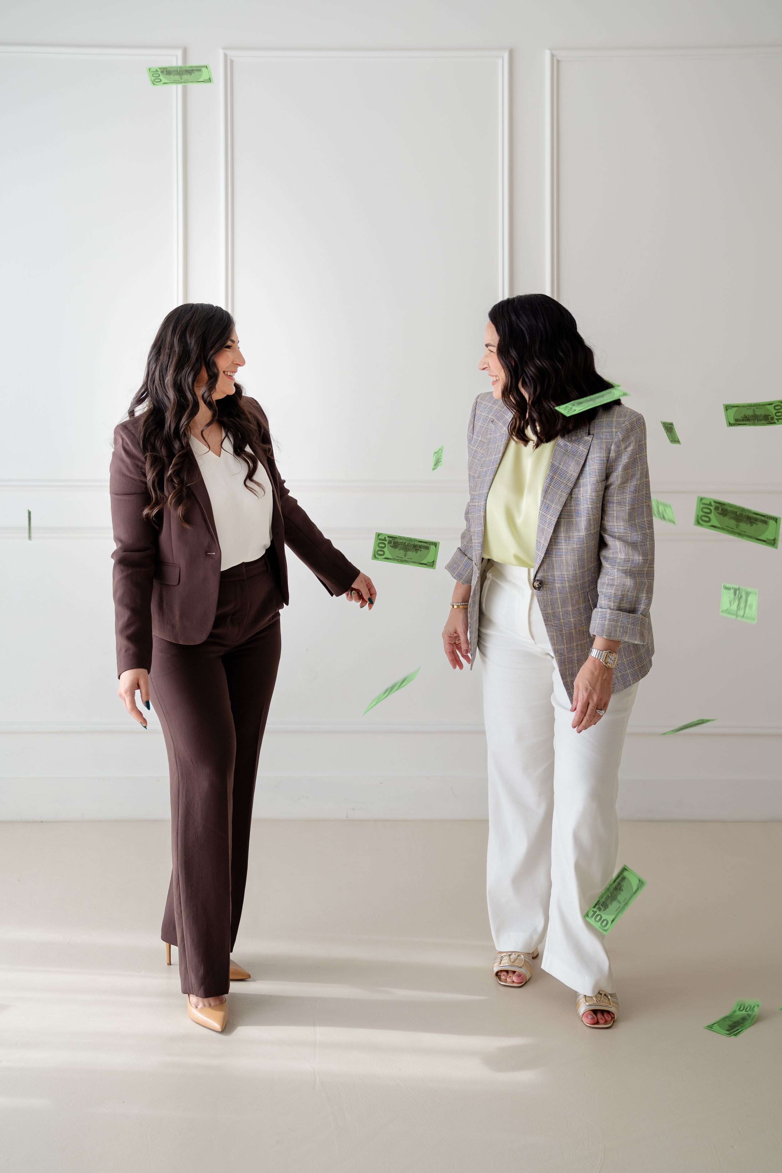 Two people in contrasting brown and white outfits interact playfully as green paper money falls around them in a bright room.