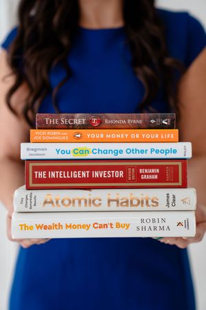 Person in blue dress holds stack of self-improvement books including The Secret, Atomic Habits and The Intelligent Investor.
