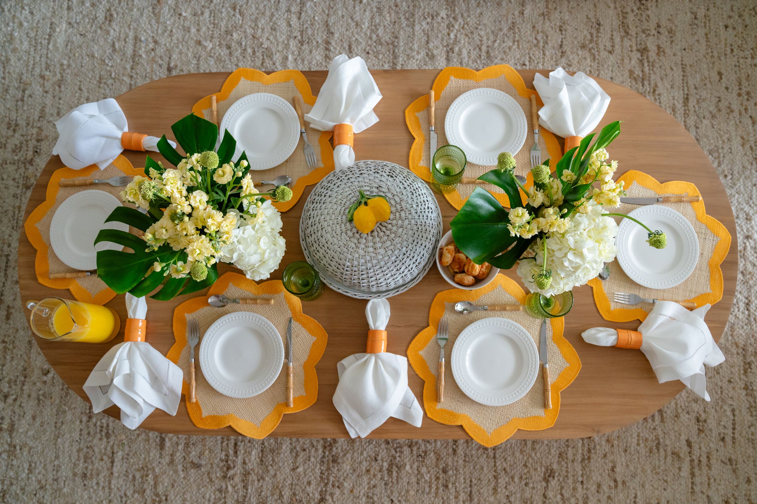 Elegant oval dining table set with yellow placemats, white plates, napkins, and fresh floral centerpieces.