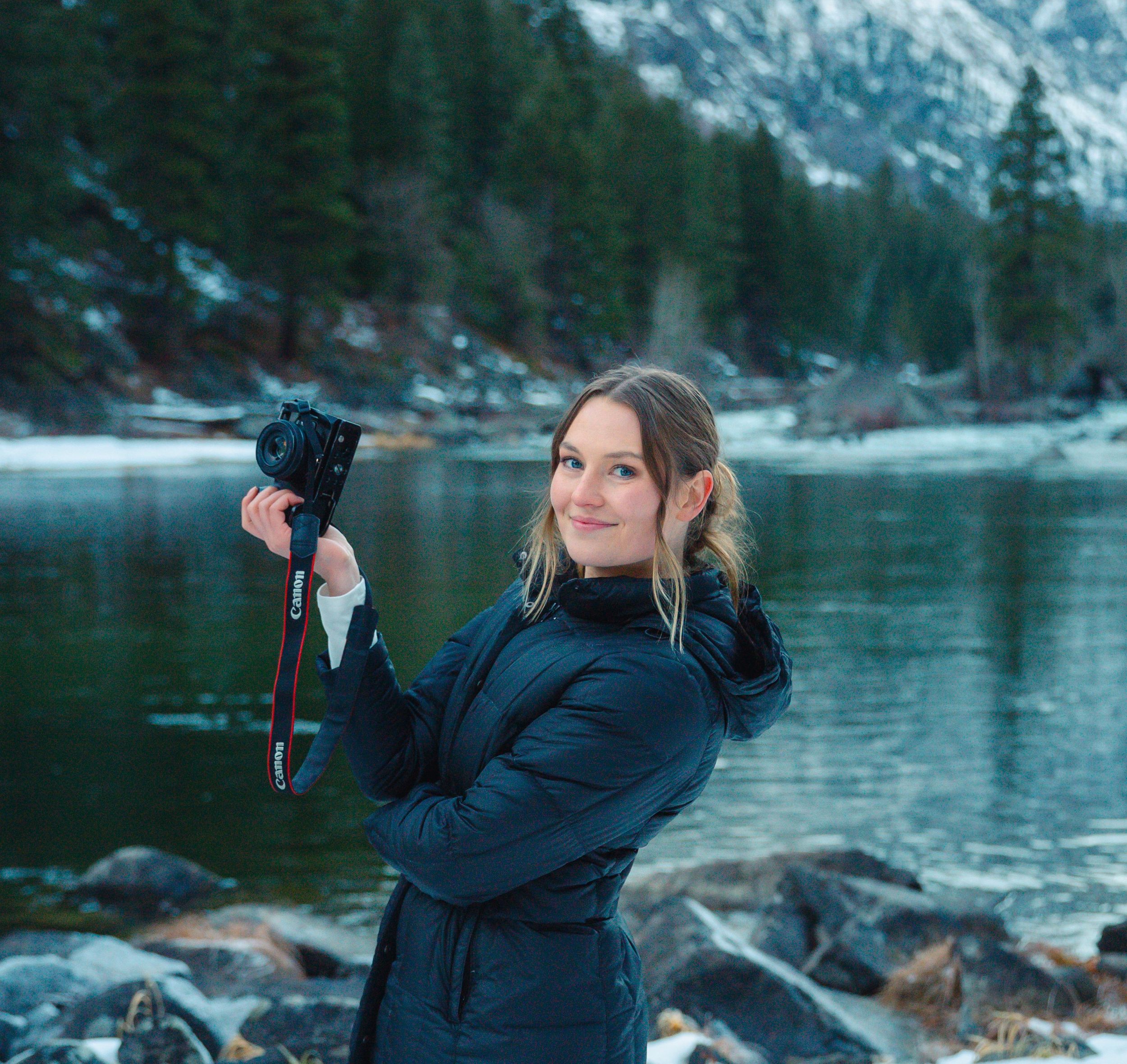A photographer wearing a winter coat poses with camera gear beside a snow-capped mountain lake.
