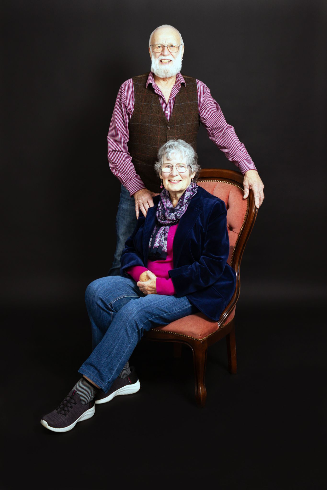 Studio portrait of two people posed on brown chair against black background.