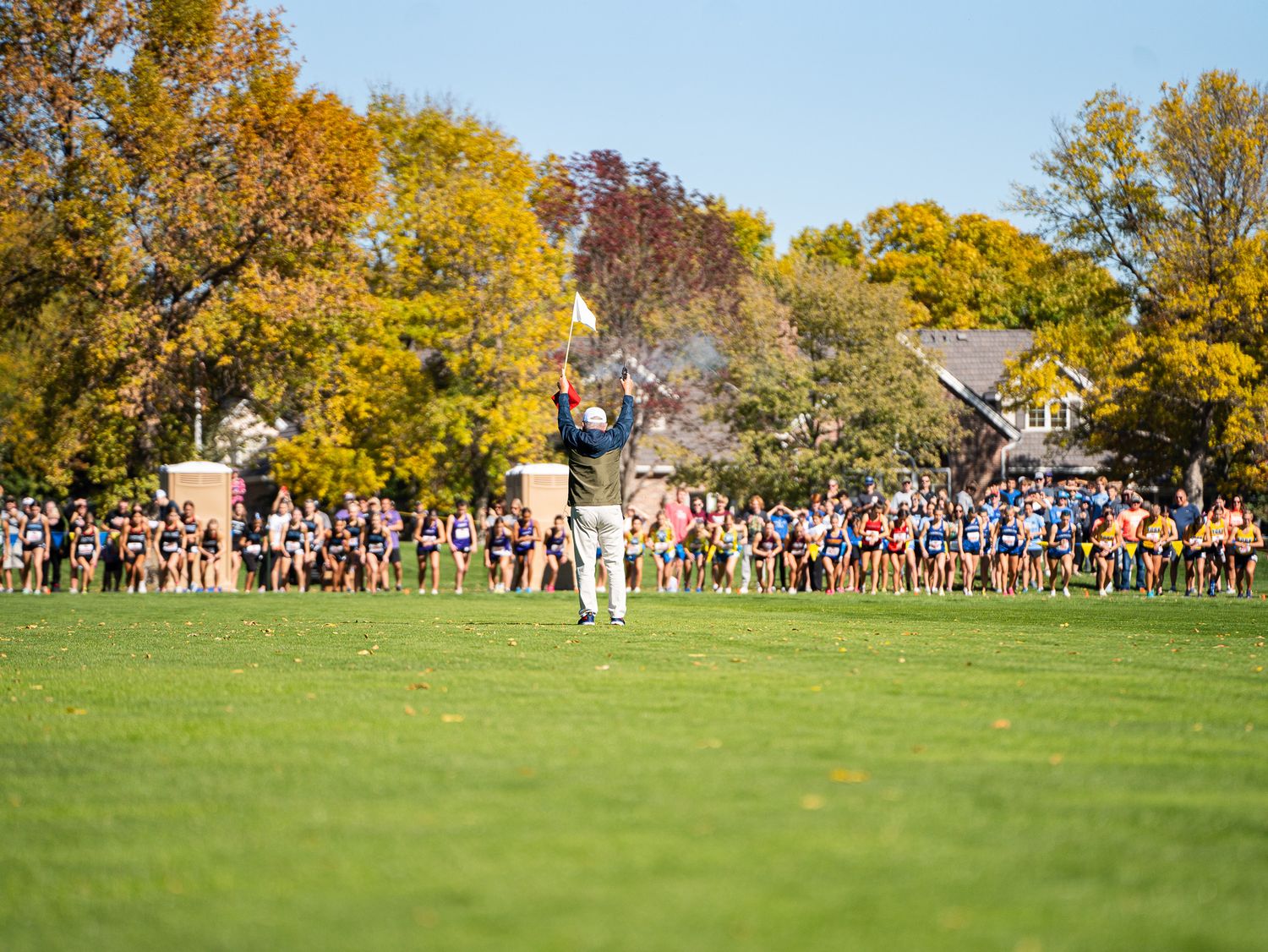 Large crowd watches cross country race on grassy field surrounded by fall foliage.