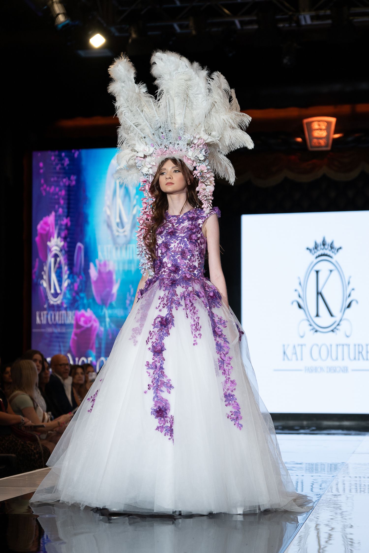 Model walks the runway in a flowing lilac midi dress with pleated skirt and bow detail at fashion show.
