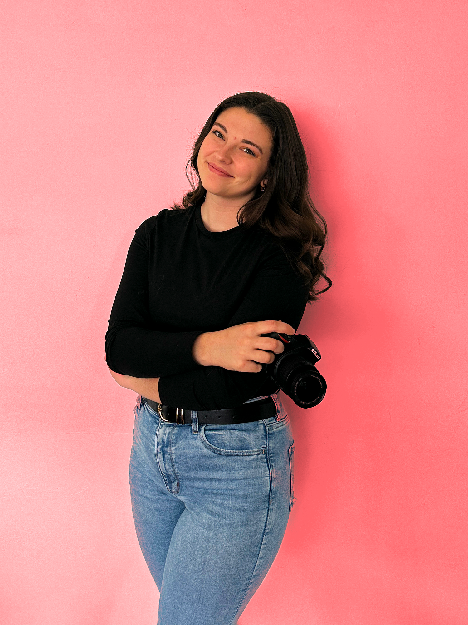 A person wearing a black sweater and blue jeans poses casually against a pink background.