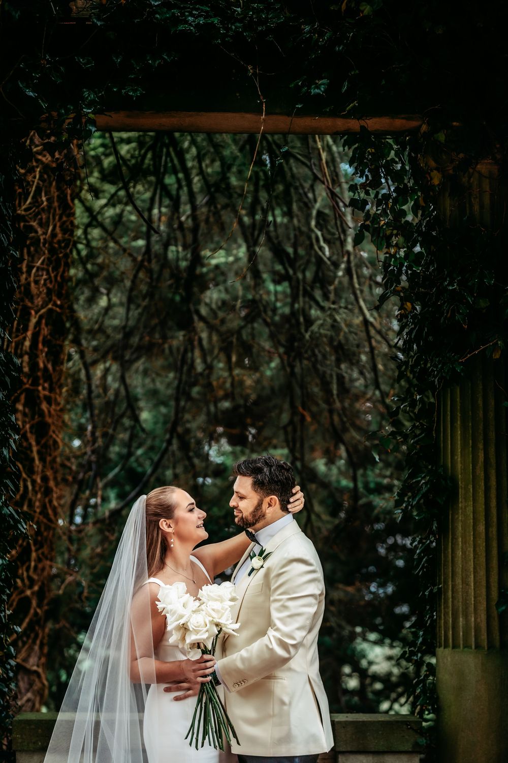 A couple in wedding attire share a romantic moment under a lush garden archway with hanging vines and greenery.