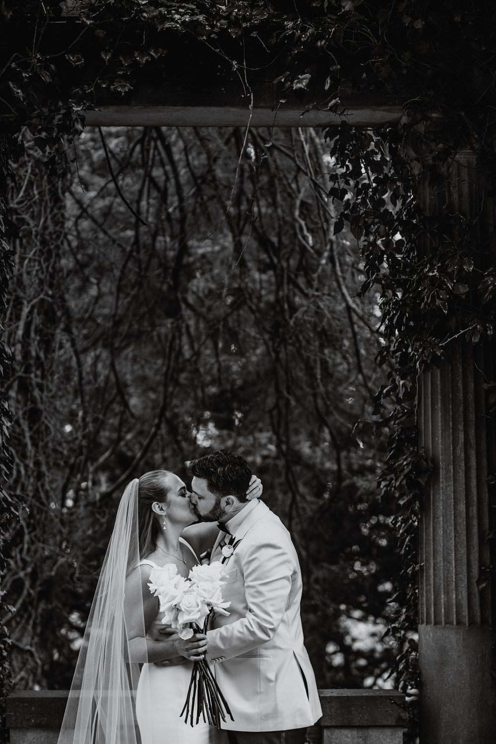 A romantic black and white wedding photo series showing a couple embracing and kissing in an outdoor garden setting.