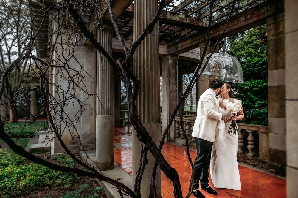 A couple in white wedding attire embraces on a brick pathway surrounded by columns and greenery.