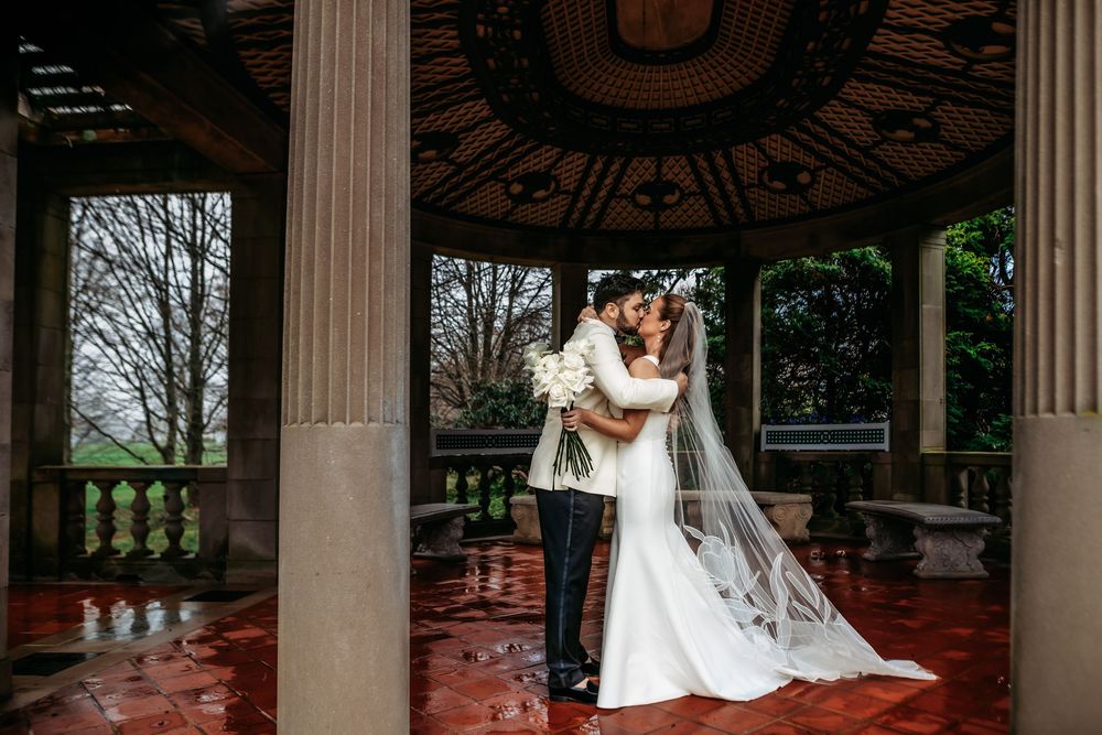 A couple embraces under an ornate gazebo with stone columns and red floor on a moody overcast day.