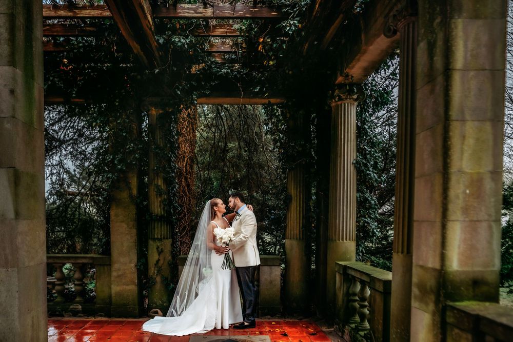 A bride and groom share an intimate moment under stone columns at a grand outdoor venue.