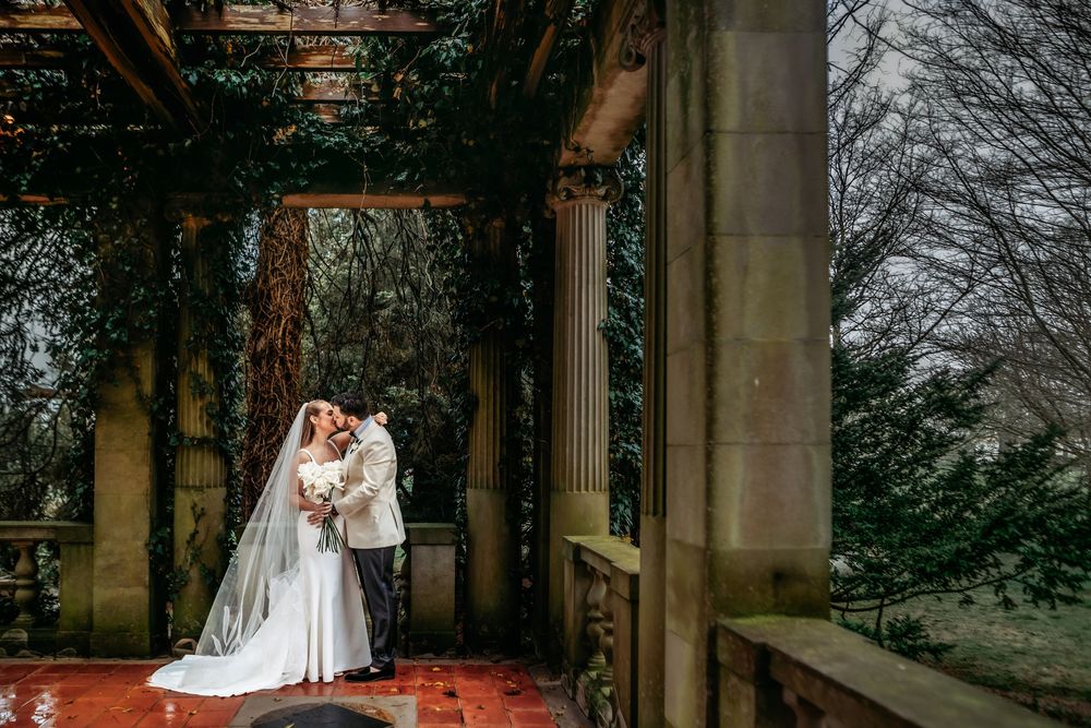 A couple in wedding attire poses under stone archways surrounded by autumn trees and foliage.