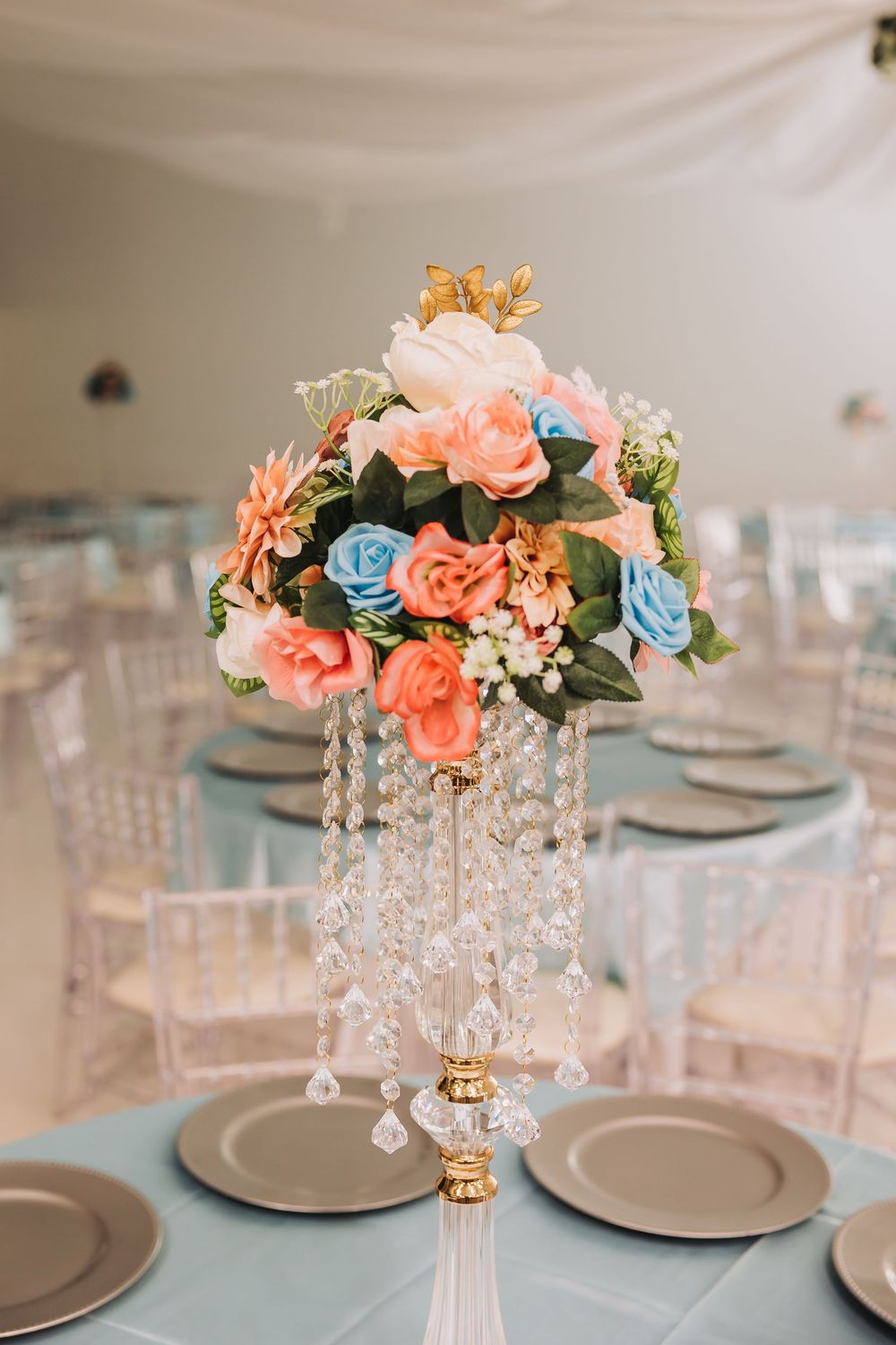 Elegant wedding centerpiece featuring coral and blue roses with crystal droplets on a gold stand at a formal table setting.