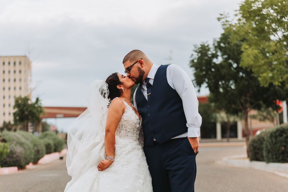 A romantic wedding couple shares a tender kiss on a sunny day with trees and buildings in the background.