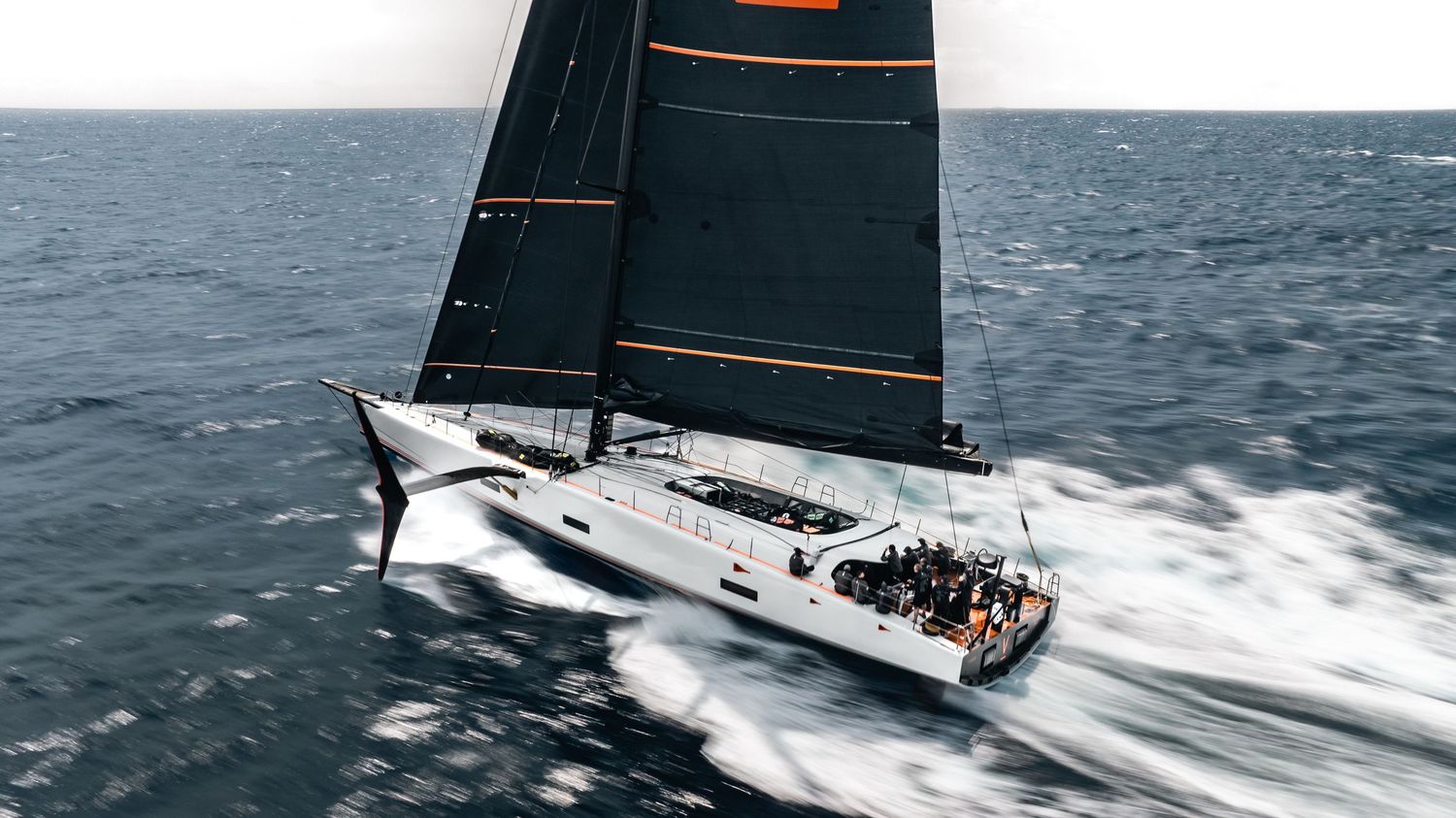 Racing sailboat cutting through ocean waves with black sails against blue sky.