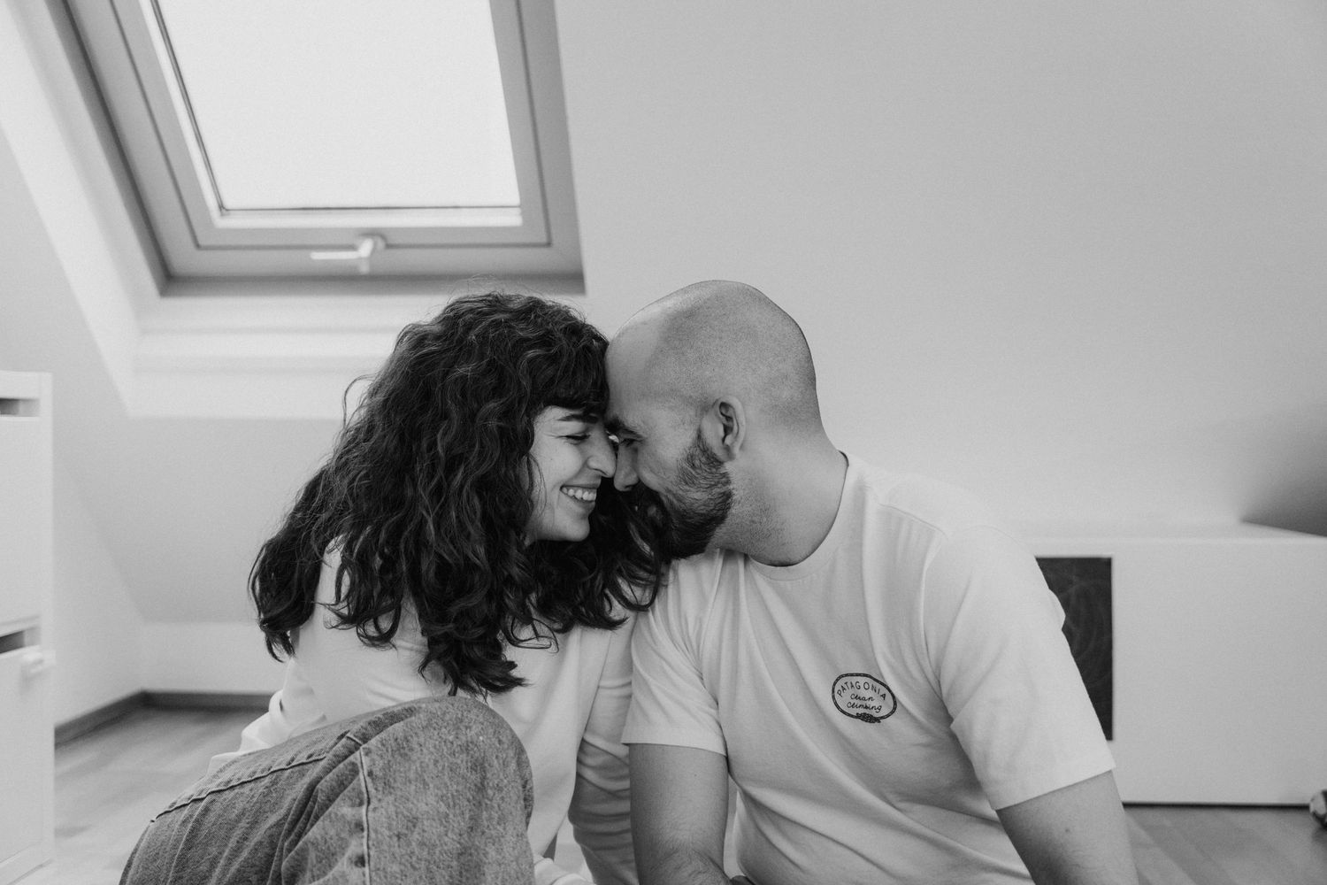 A tender black and white intimate moment captured between a couple touching foreheads in an attic room.