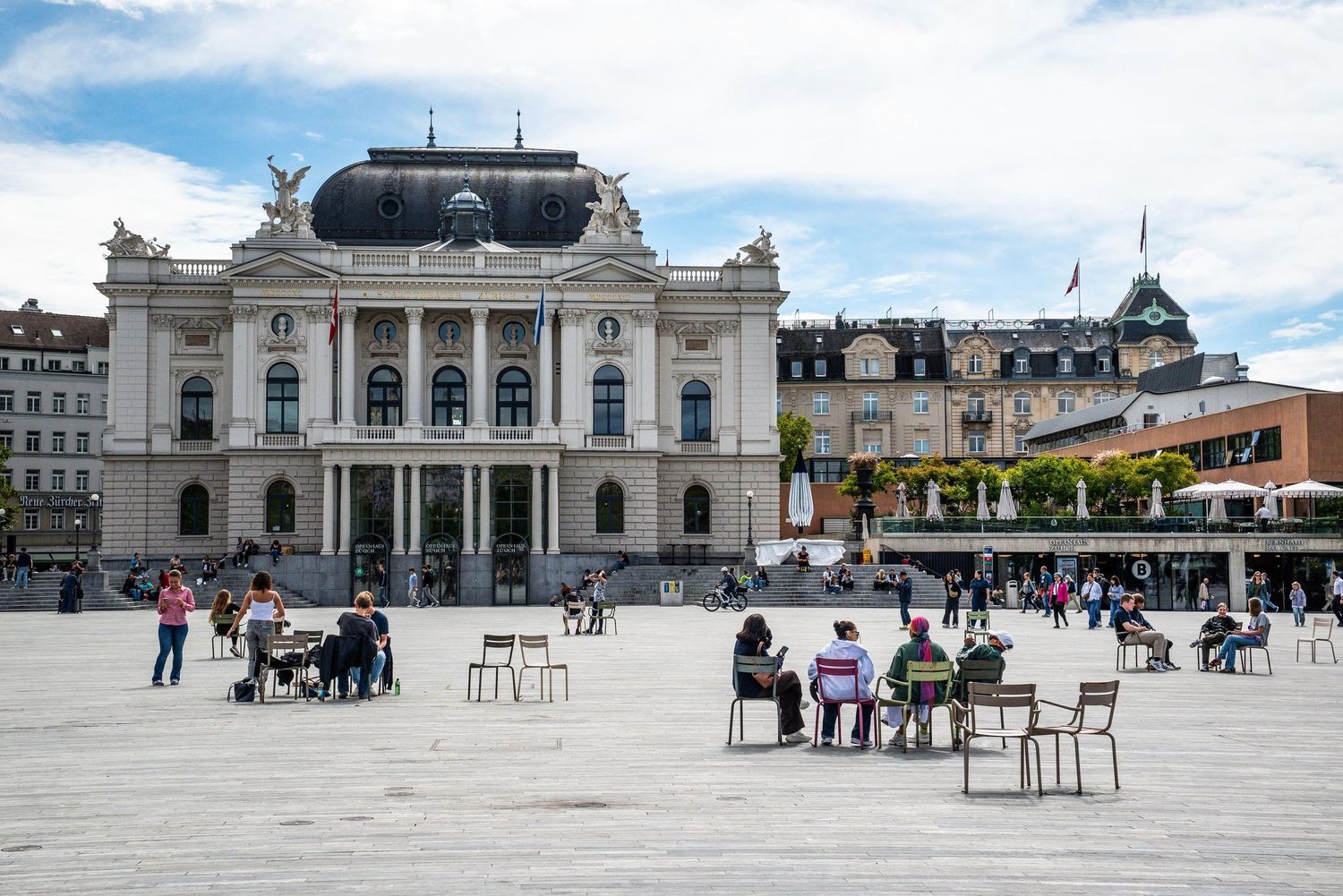 Panoramic view of a historic European plaza with classical architecture and scattered chairs under a cloudy sky.