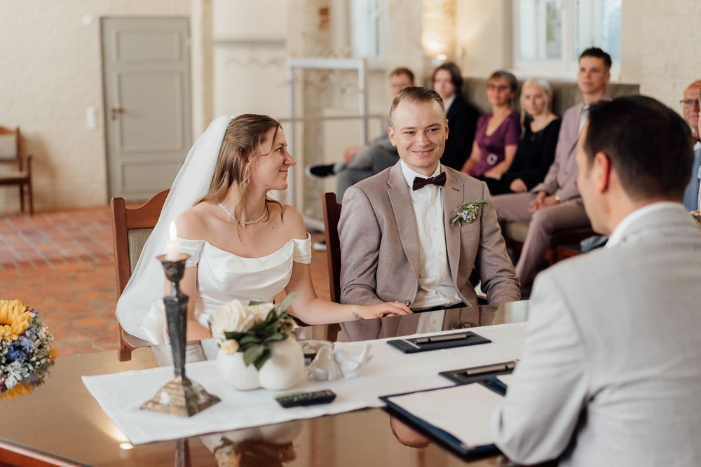 Bride and groom sign marriage documents at a bright indoor ceremony while guests look on from their seats.